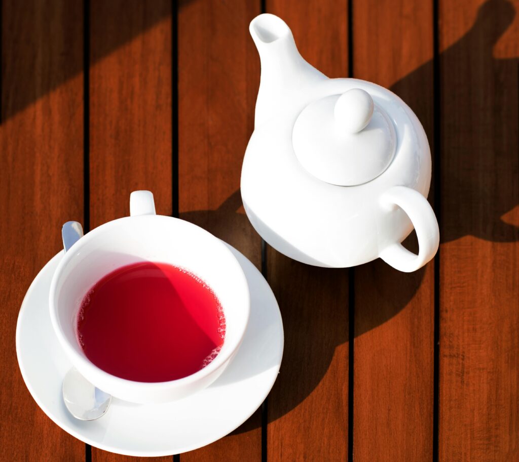 A clear glass cup filled with bright red hibiscus tea, surrounded by dried hibiscus petals on a wooden table.