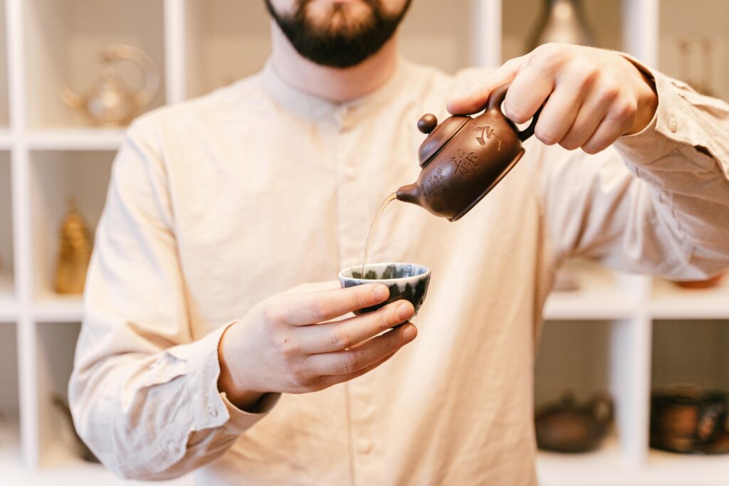 Close-up of a man pouring tea from a traditional brown teapot into a small cup indoors.