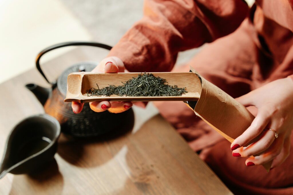 A close-up view of hands handling loose tea leaves with a wooden scoop alongside a teapot.