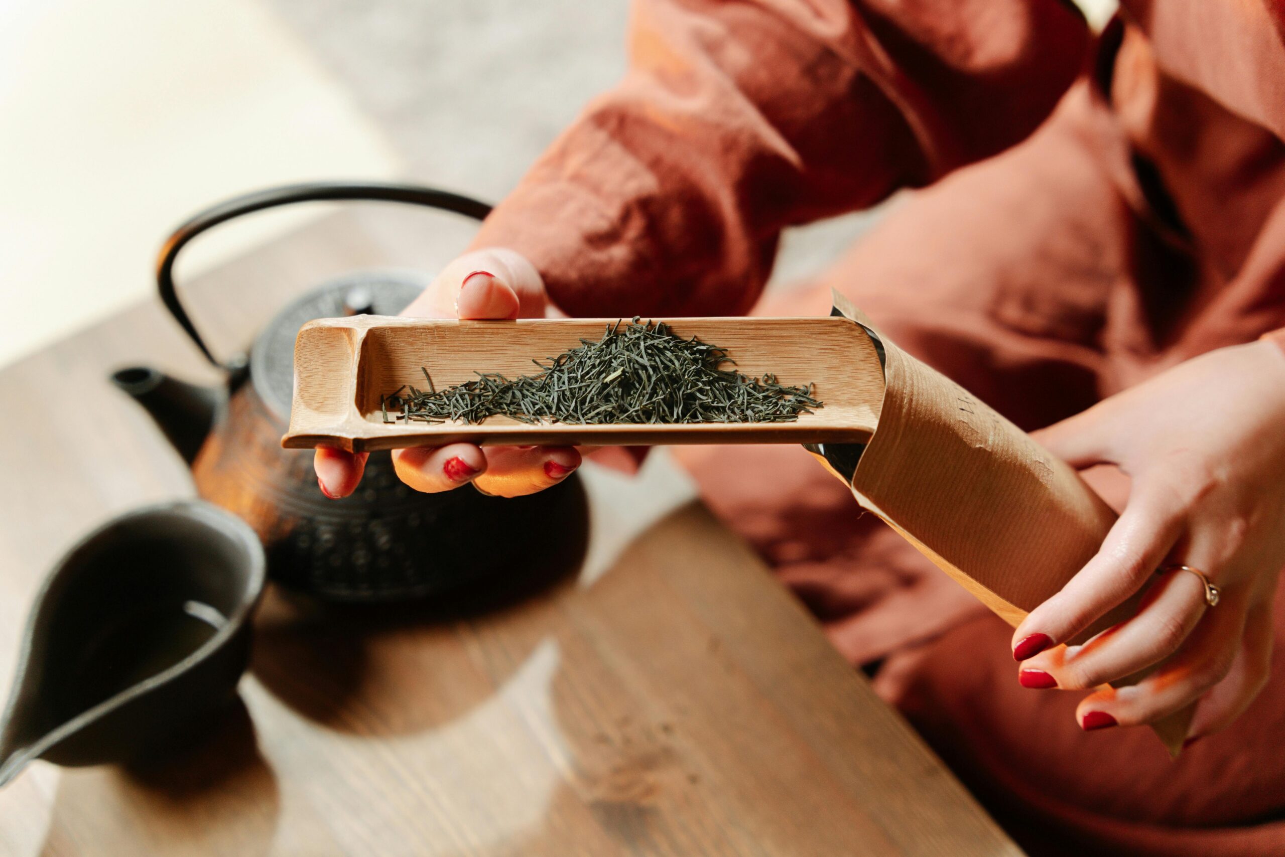 A close-up view of hands handling loose tea leaves with a wooden scoop alongside a teapot.